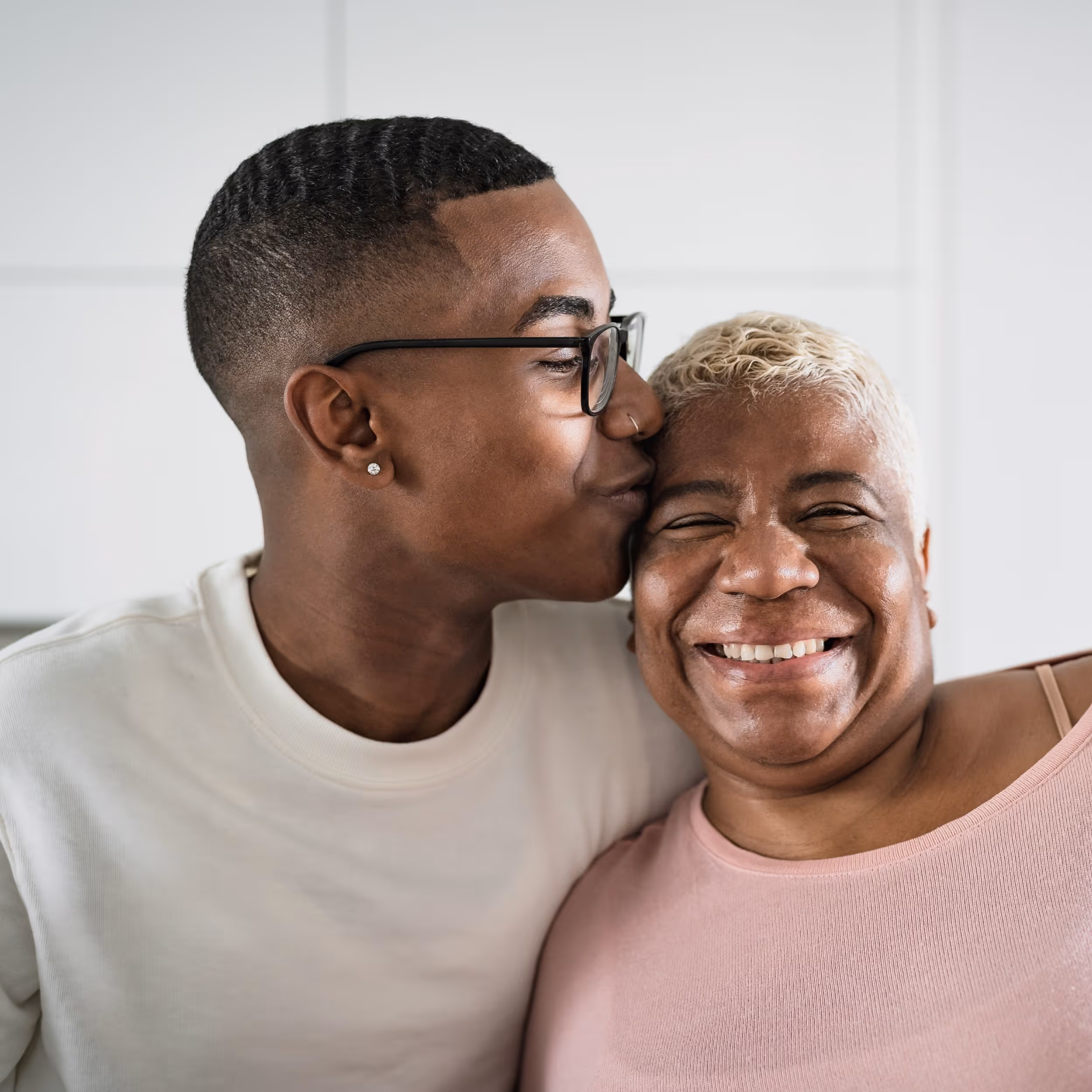 A young man wearing glasses and a white t-shirt kisses the forehead of his mum, who smiles widely