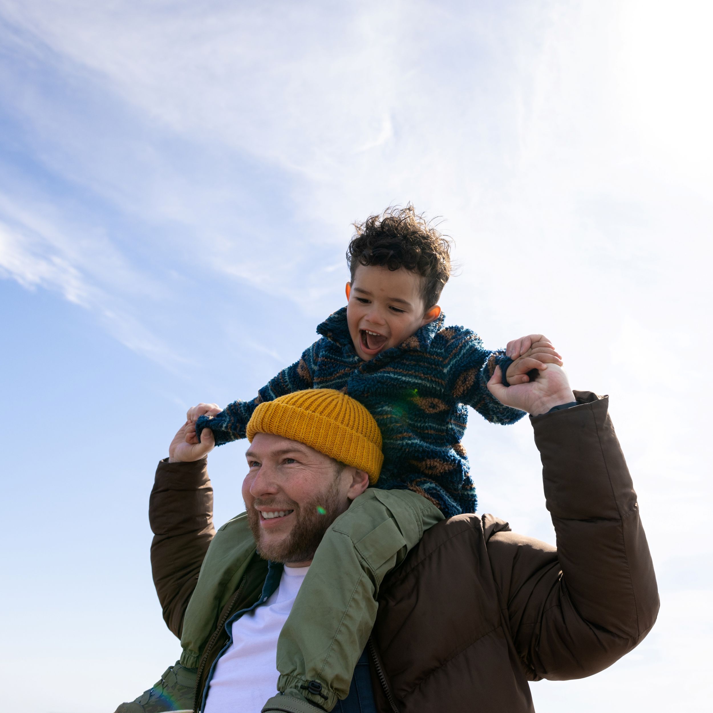A man in a yellow woollen hat carries a small boy on his shoulders, against a backdrop of blue sky streaked with clouds. 