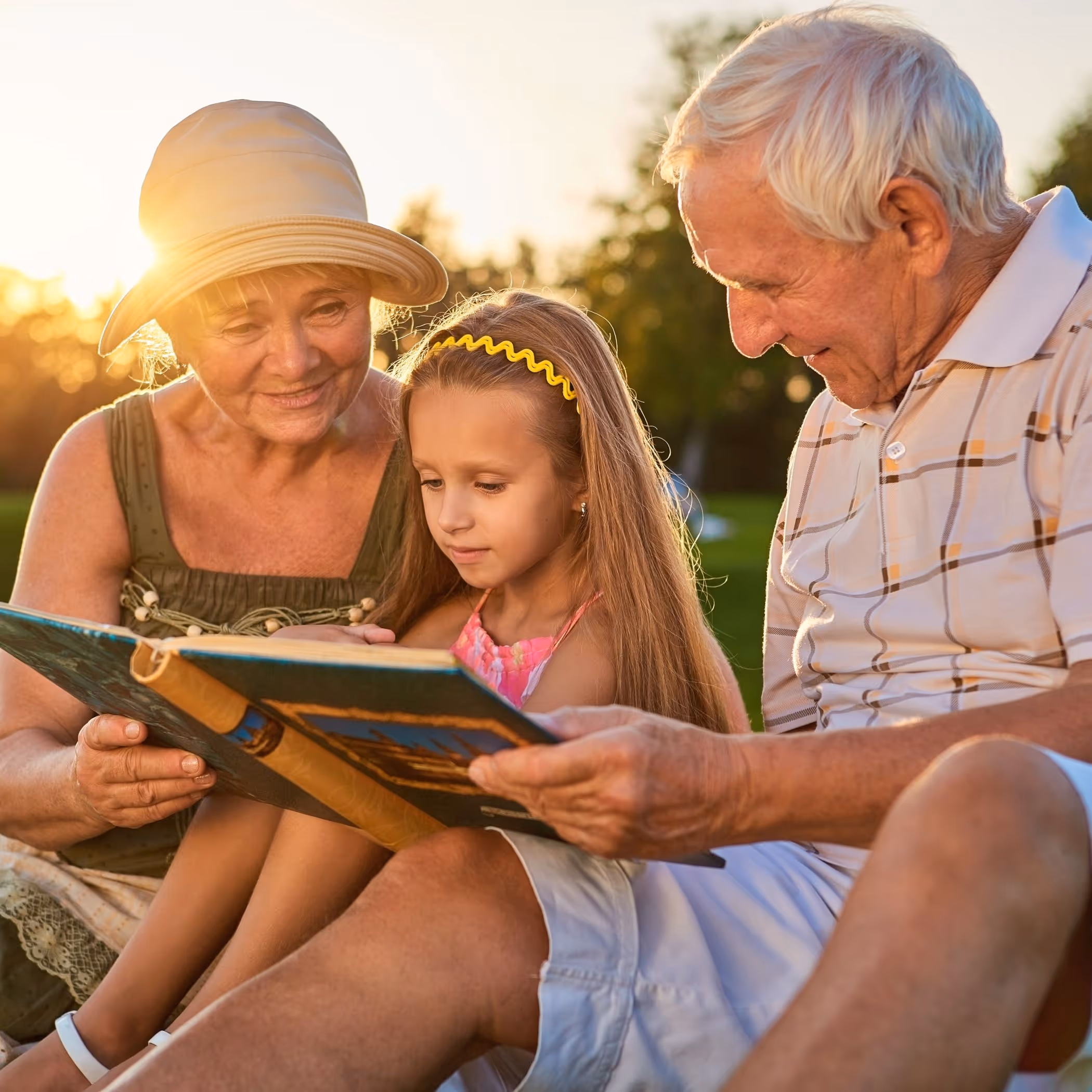 Older couple reading a book with their small granddaughter, sitting outside on a sunny lawn.