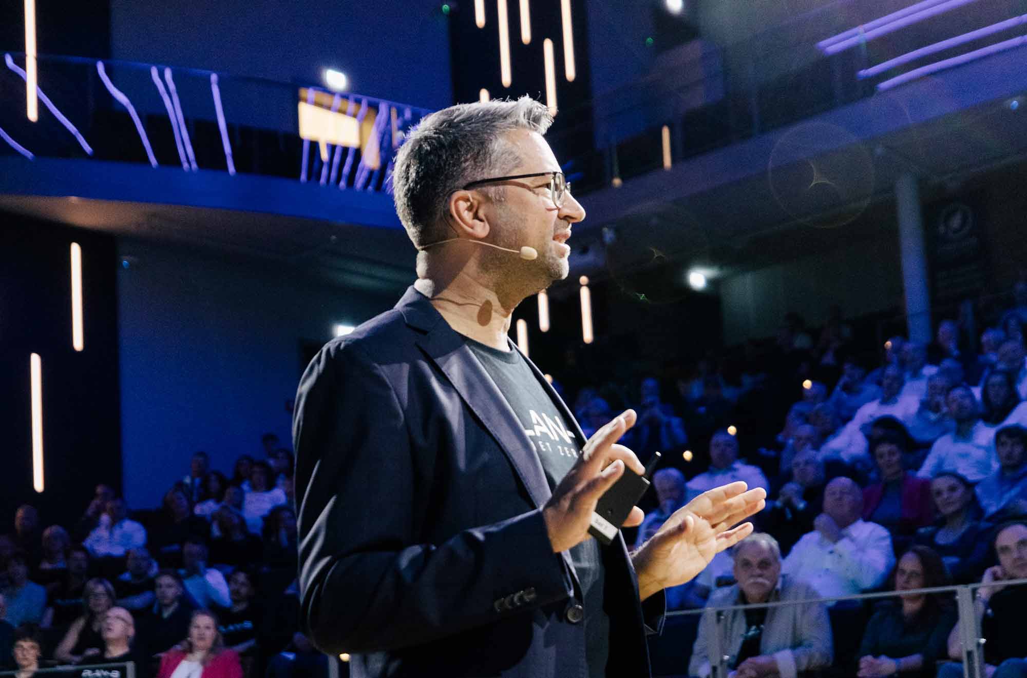 Ein junger Mann in weißem Hemd steht lächelnd an einem Konferenztisch und bespricht mit mehreren Kolleg:innen ein Projekt. Die Gruppe wirkt engagiert und gut gelaunt. Auf dem Tisch liegen Dokumente, im Hintergrund sind Computerbildschirme mit Diagrammen zu sehen. Die Szene spielt in einem modernen Büro mit Backsteinwand und großen Fenstern.
