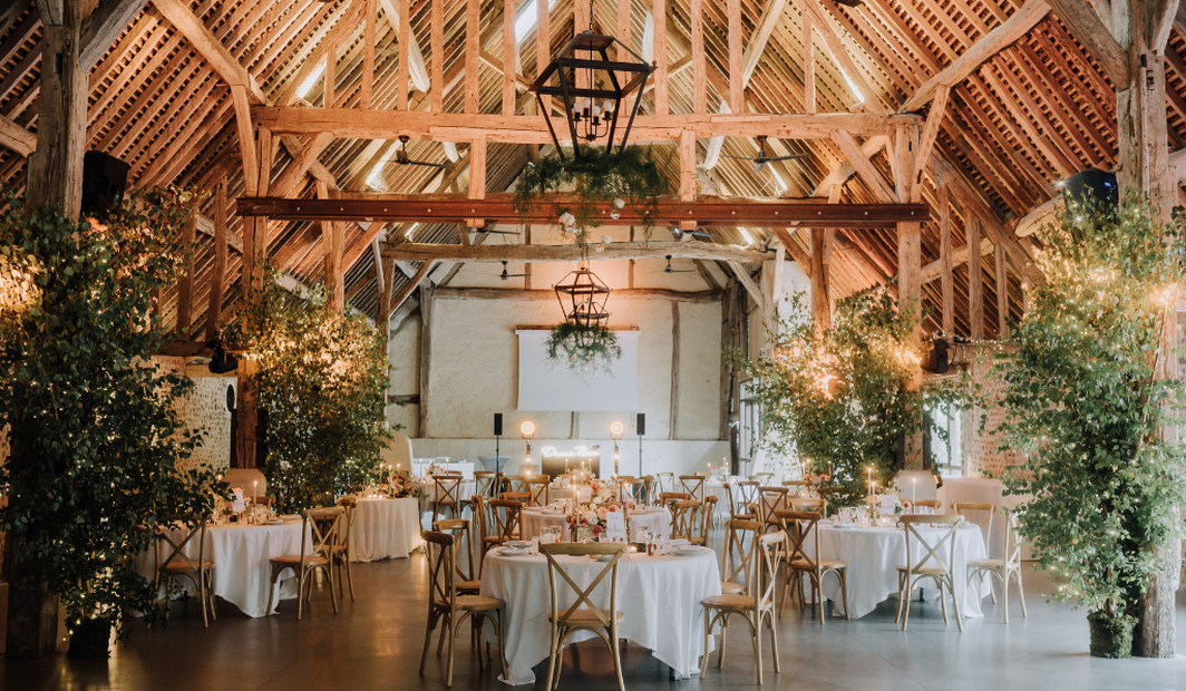 Salle de réception en grange rénovée, décorée de guirlandes lumineuses et de verdure pour un dîner de mariage.