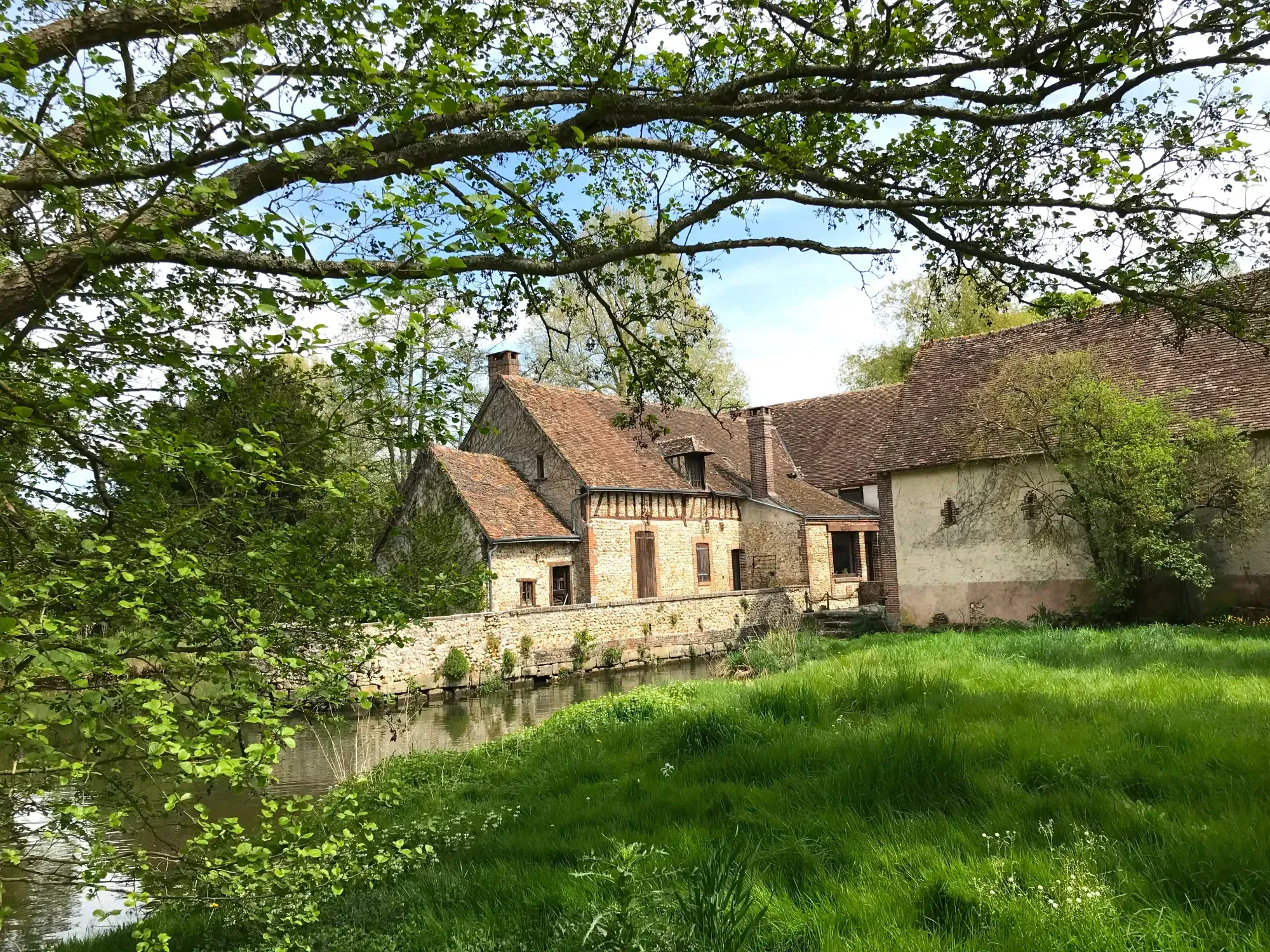 Old stone house by a calm river, surrounded by greenery and trees under a blue sky, in a traditional watermill style.