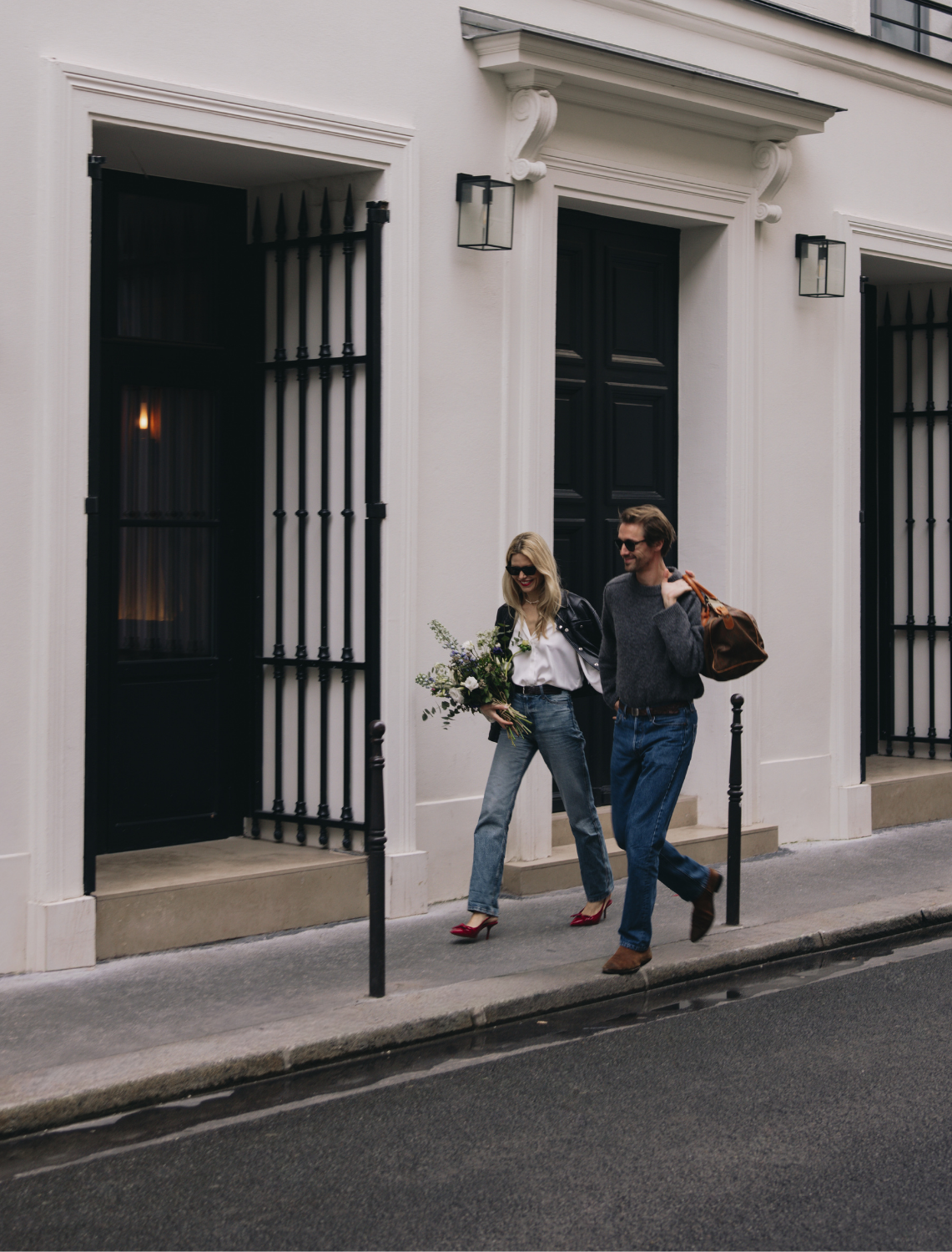Un couple marche sur un trottoir devant un bâtiment blanc avec une femme portant un bouquet de fleurs.