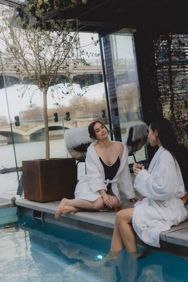 Two women in white robes relaxing by an indoor pool, one with feet in water, holding drinks.