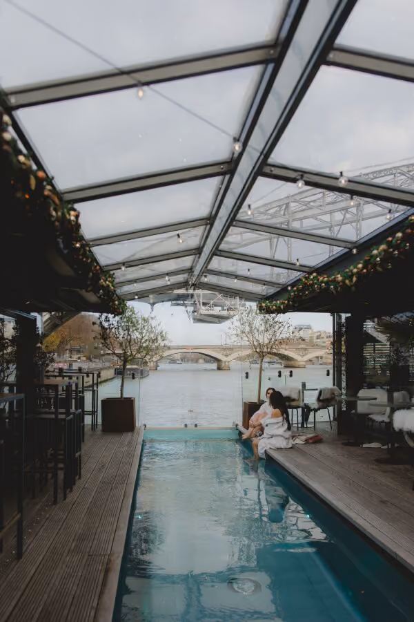 Covered outdoor pool with two people sitting on edge, overlooking a river and bridge in the background.