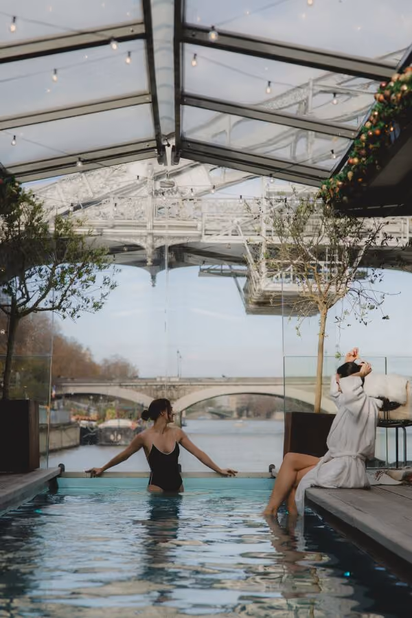 Woman in black swimsuit sits in pool while another in robe sits on edge near glass wall with bridge view.