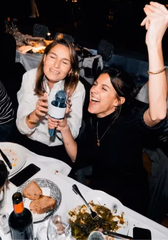 Two women laughing and singing together at a dinner table with food and drinks.