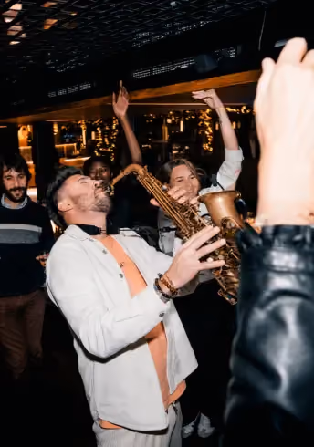 Man passionately playing saxophone with friends cheering in lively indoor setting at night.