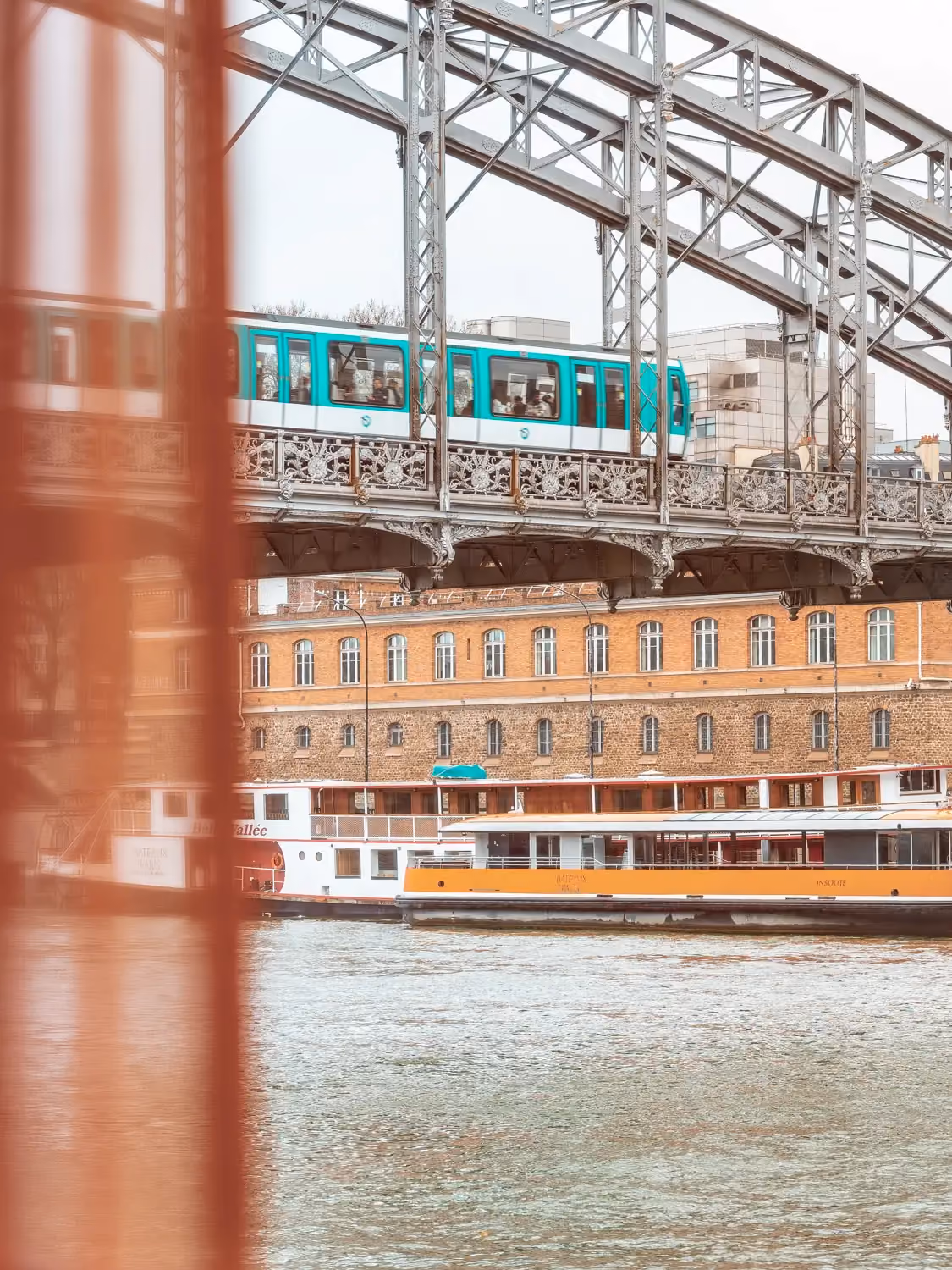 A turquoise metro train crosses a steel bridge over a river with boats and buildings behind.