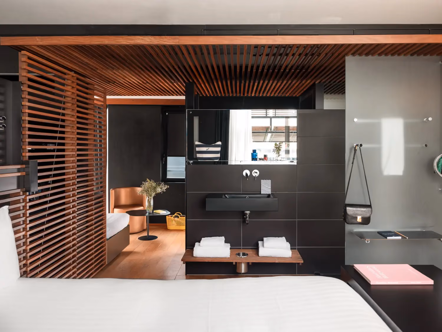 Modern bedroom with white bed, wood slat divider, black sink area, and brown chair in background.
