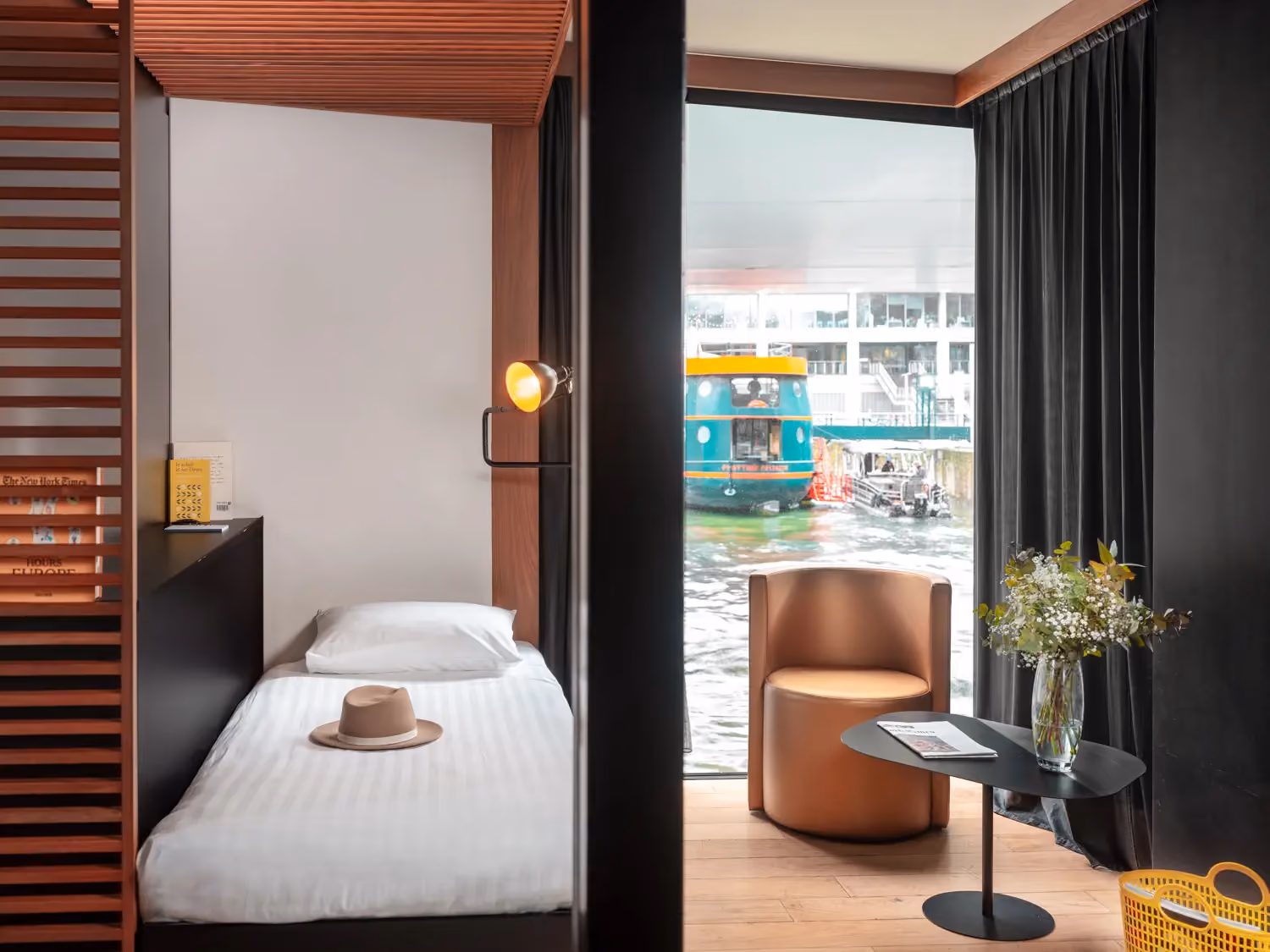 Modern hotel room with single bed, lamp, chair, small table with flowers, and waterfront view with boat.