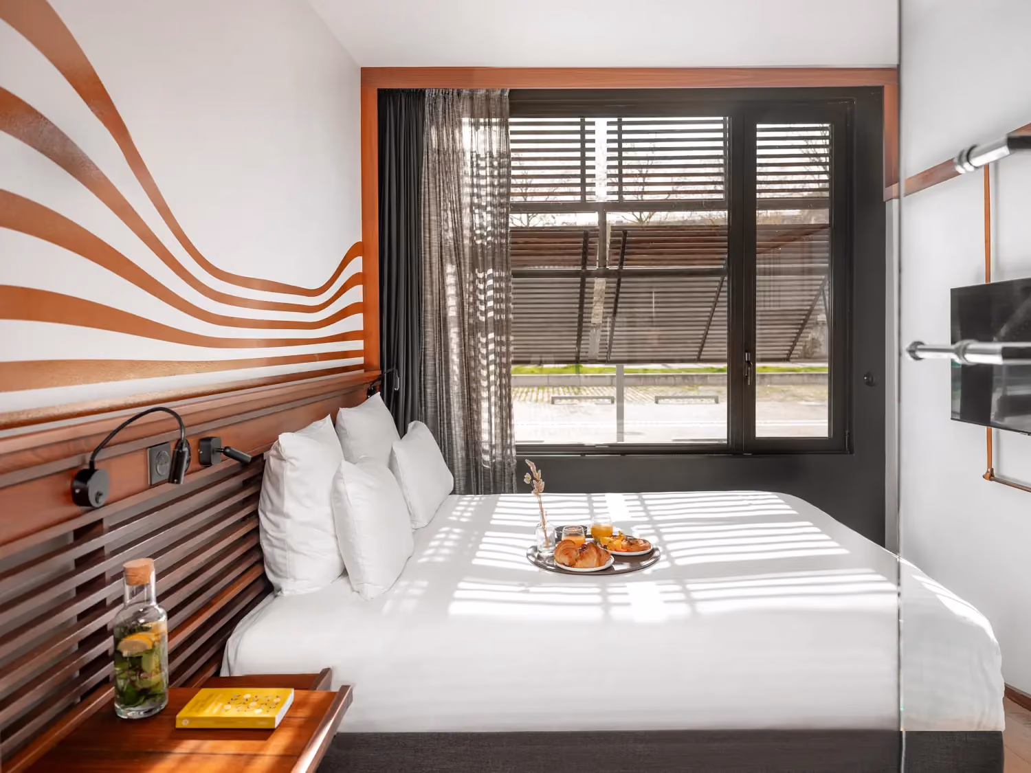 Modern hotel room with king bed, white pillows, wooden headboard, tray with croissants, and large window.