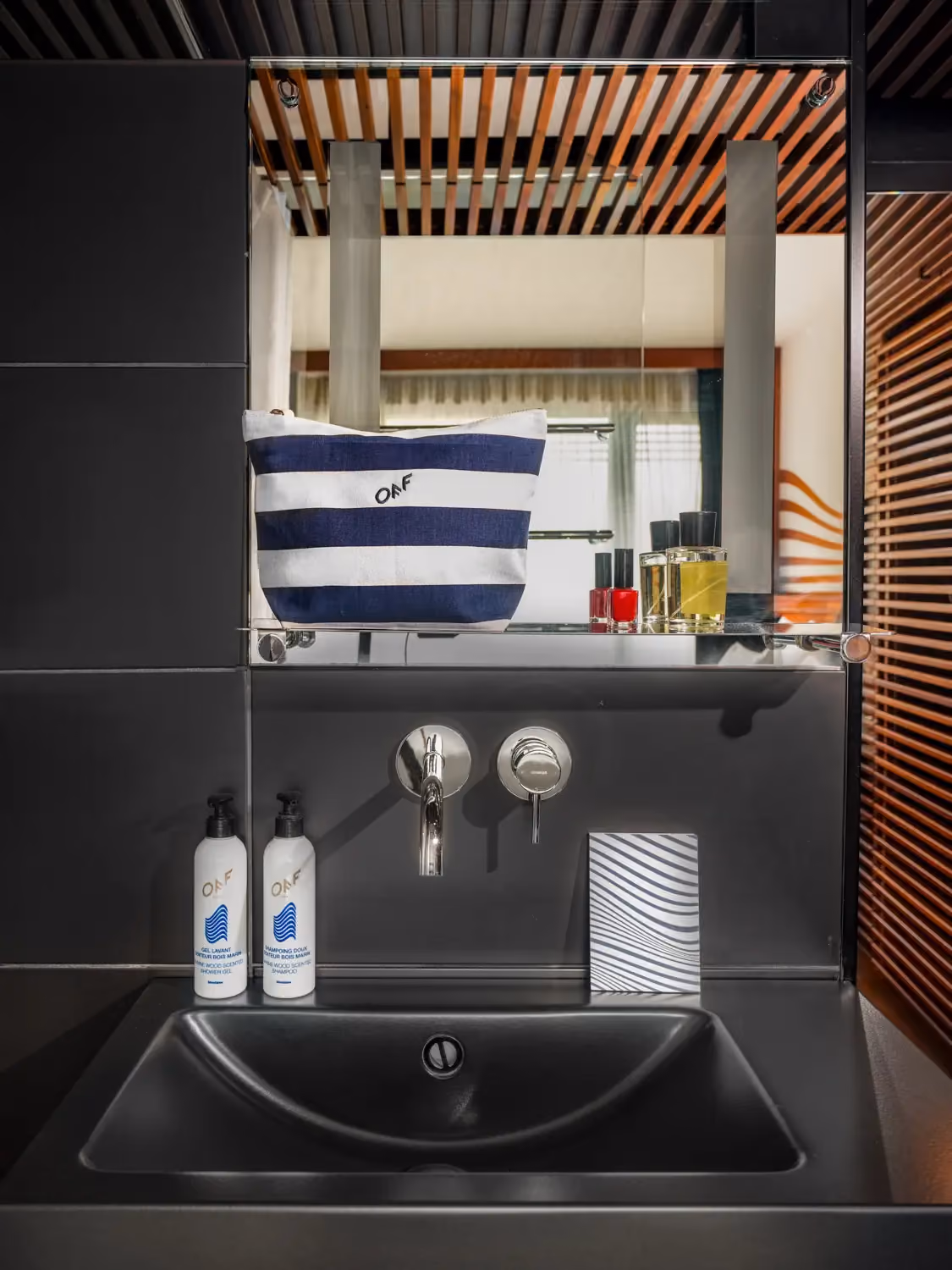 Black bathroom sink with wall-mounted faucet, two pump bottles, striped pouch, and toiletries on glass shelf.
