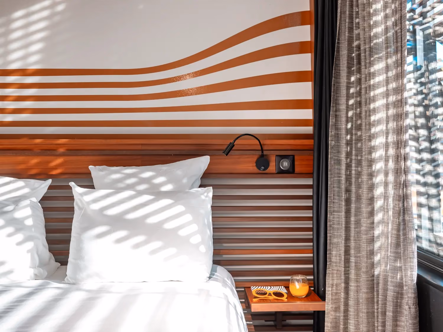 Sunlit bed with white pillows, wooden headboard, wall lamp, orange sunglasses, and orange juice glass.