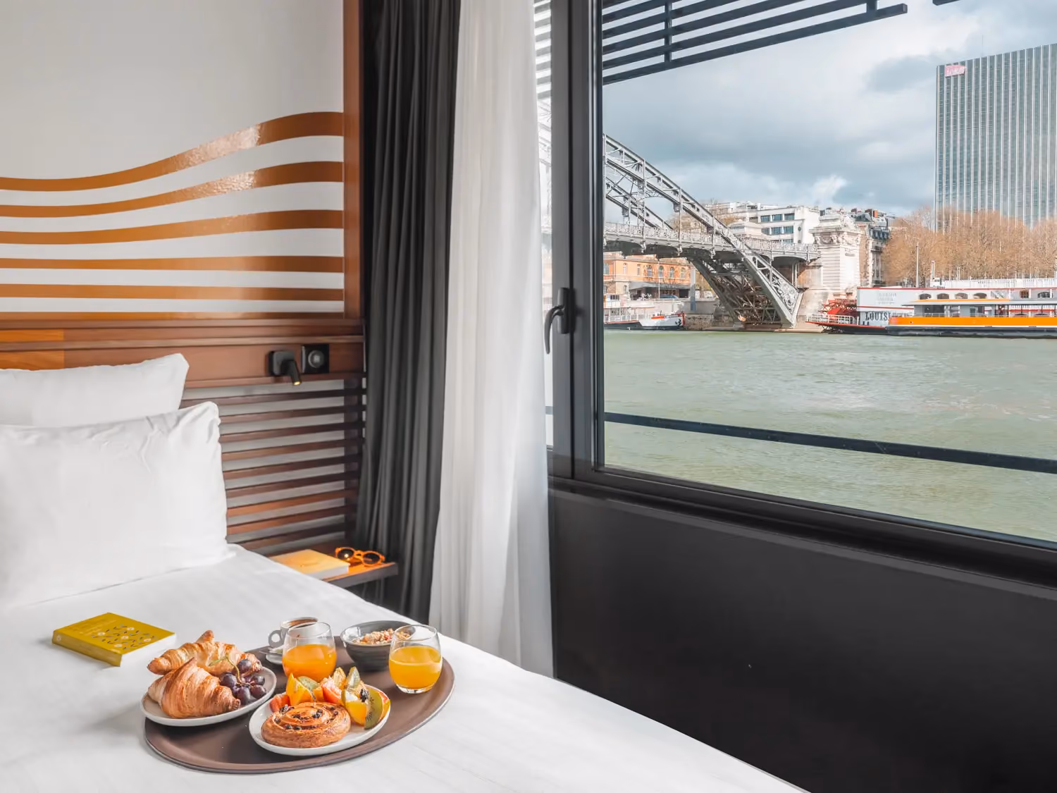 Tray with croissants, fruit, juice, and coffee on a bed by a window overlooking a river and bridge.