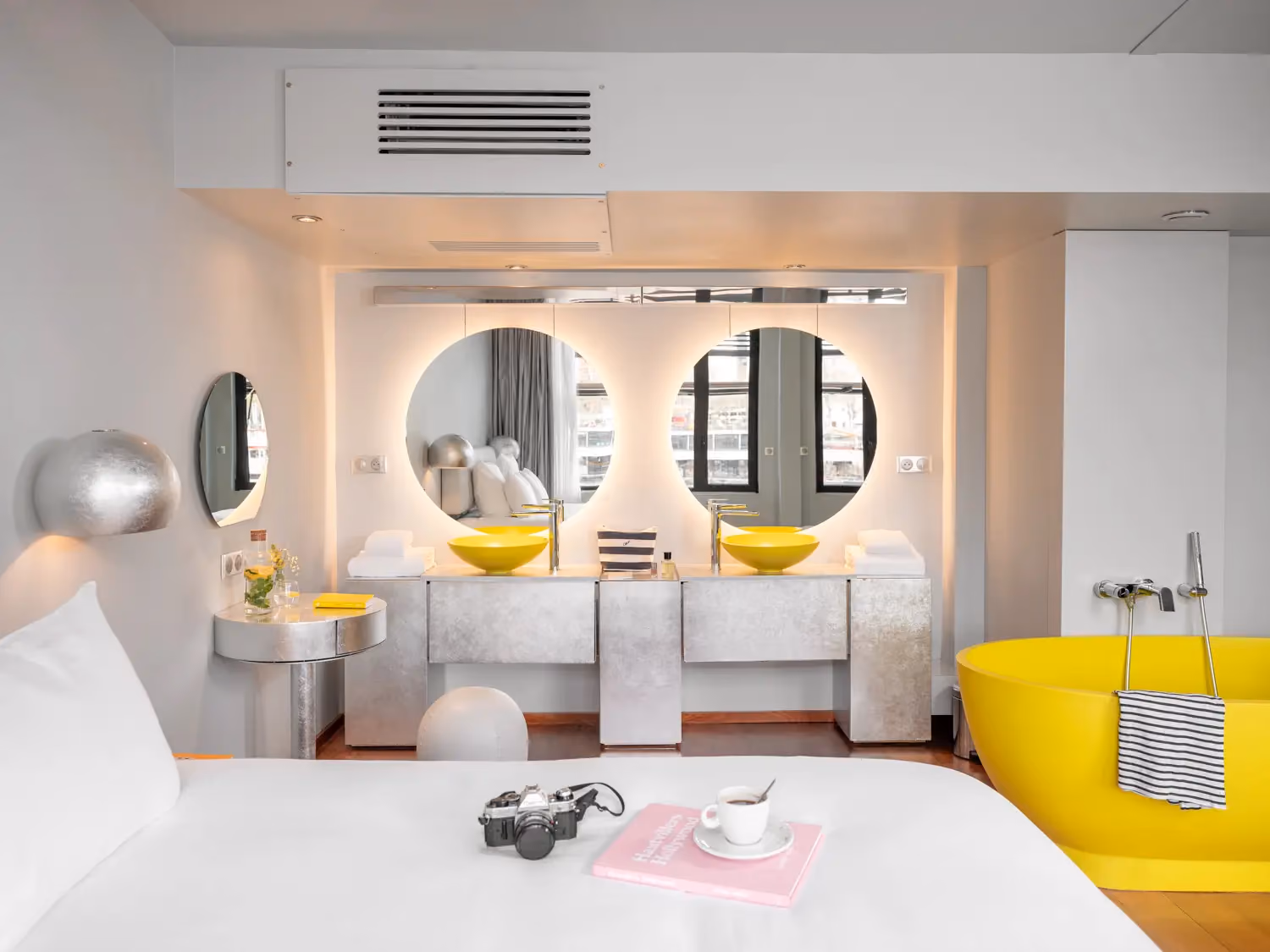 Modern bedroom with white bed, yellow bathtub, yellow sinks, round mirrors, and silver accents.