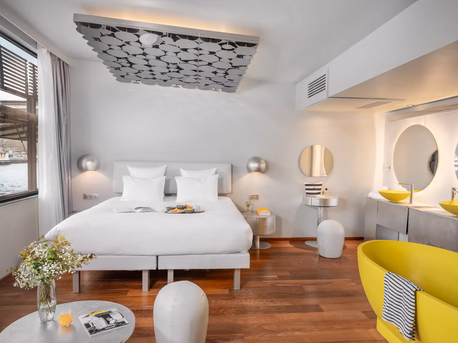 Modern bedroom with white bed, silver lamps, wooden floor, and a bright yellow bathtub with towel.