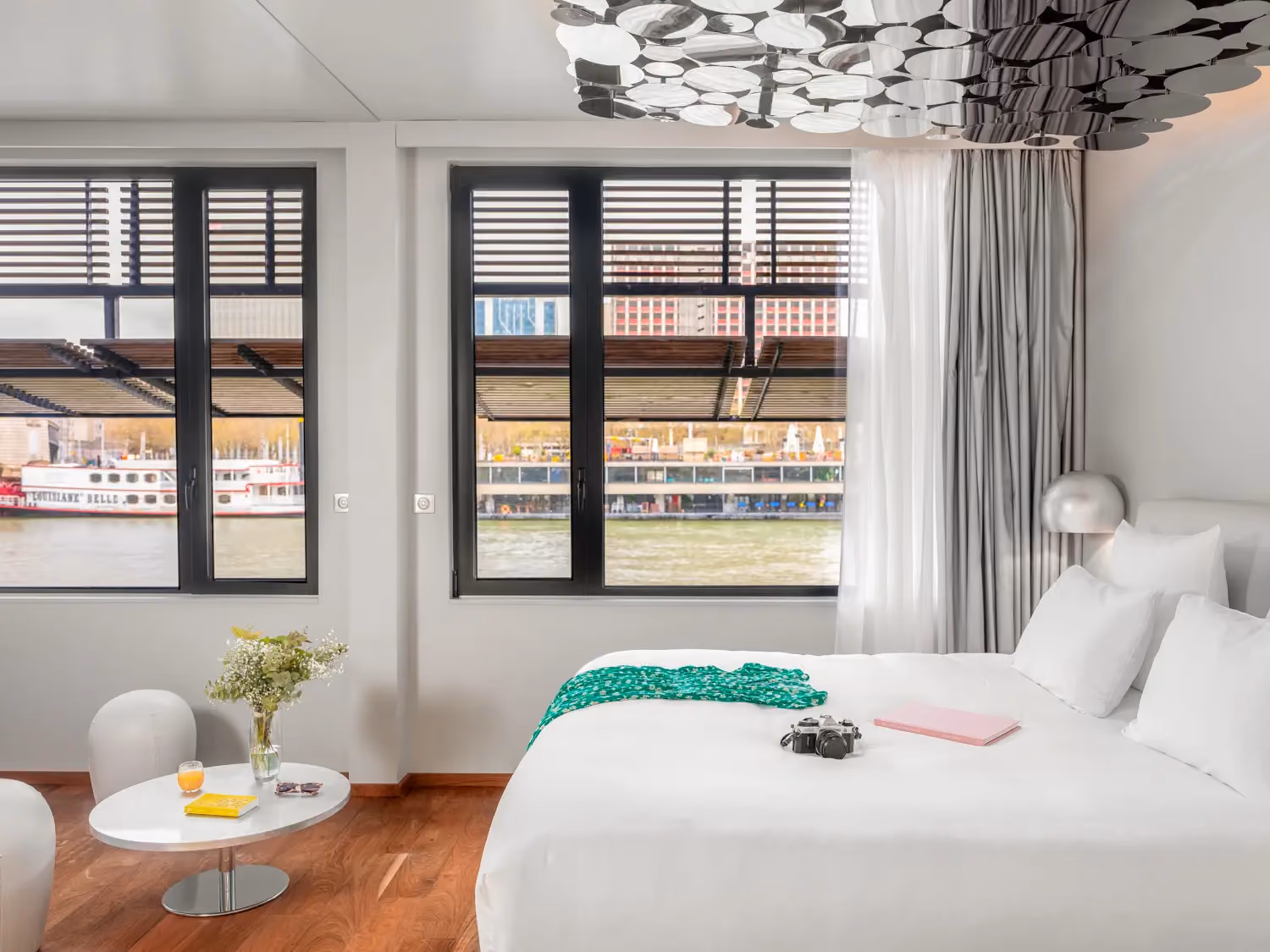 Modern hotel room with white bed, green scarf, camera, small table, and river view through two windows.
