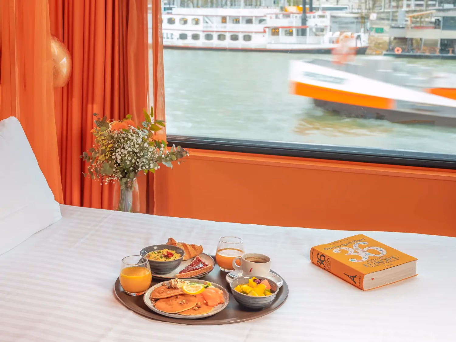 Breakfast tray with pancakes, fruit, juice, coffee, and pastries on a bed by window overlooking river boats.