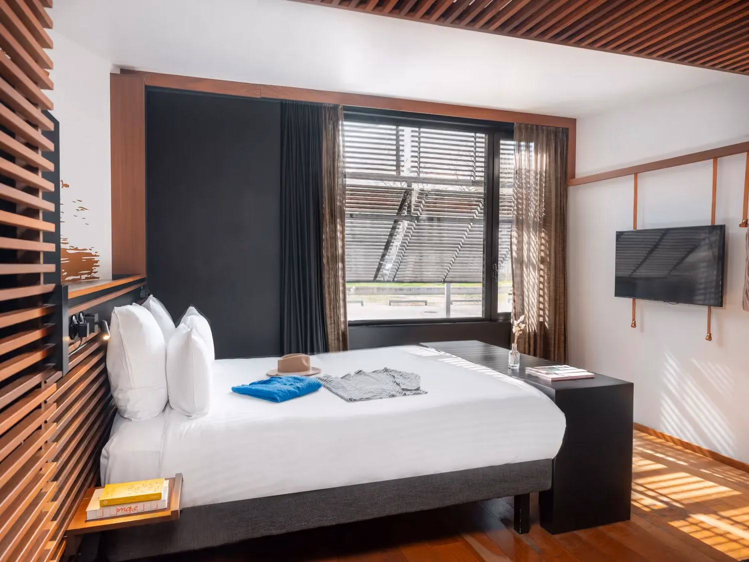 Modern bedroom with neatly made bed, wooden accents, large window with blinds, and wall-mounted TV.