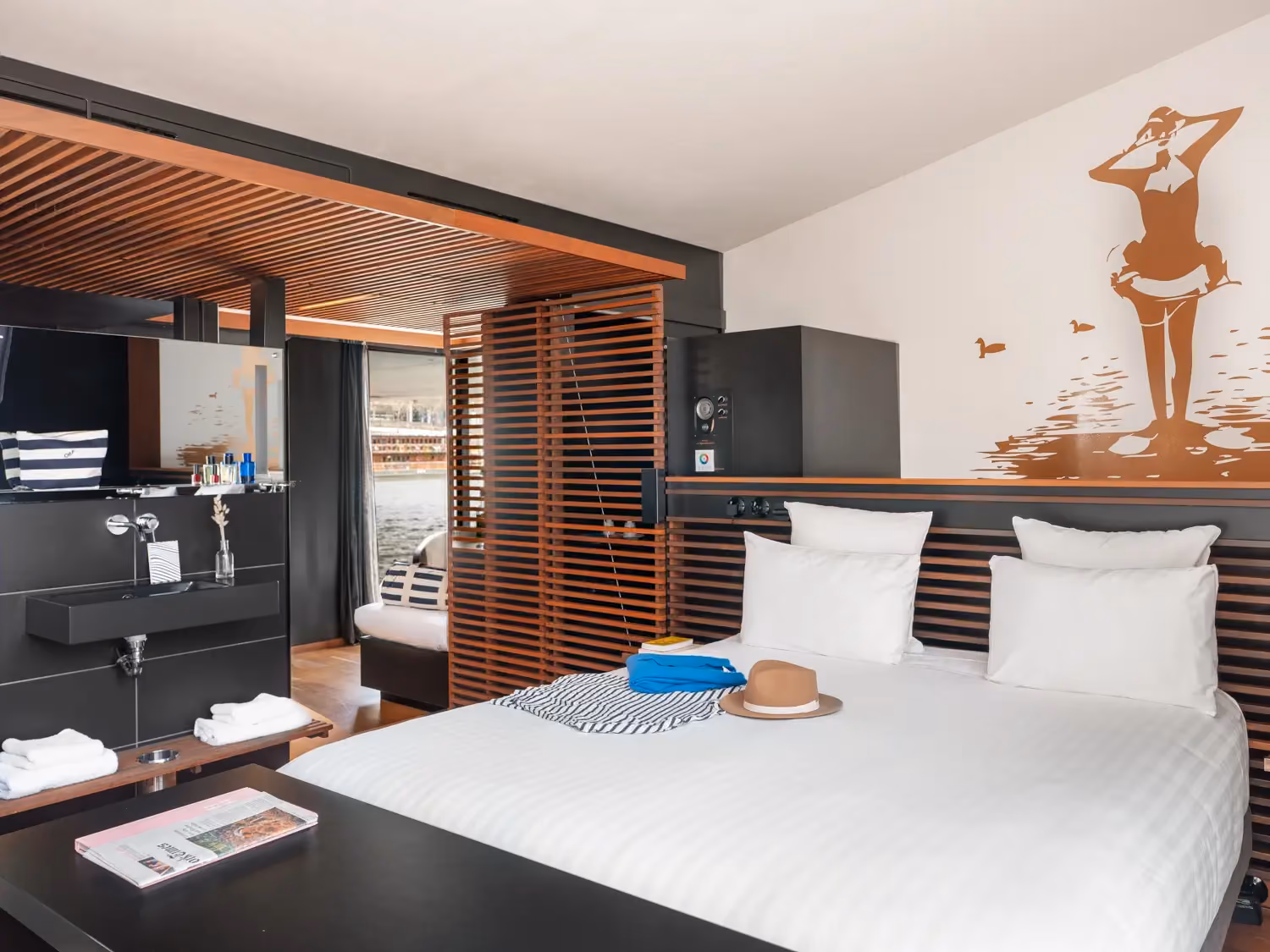Modern bedroom with white bed, striped wall art, hat on bed, wooden panel decor, and a view of water outside.