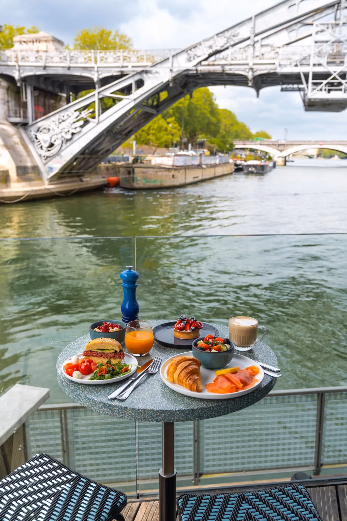 Table with breakfast dishes, juice, coffee by river with metal bridge and green trees in background.
