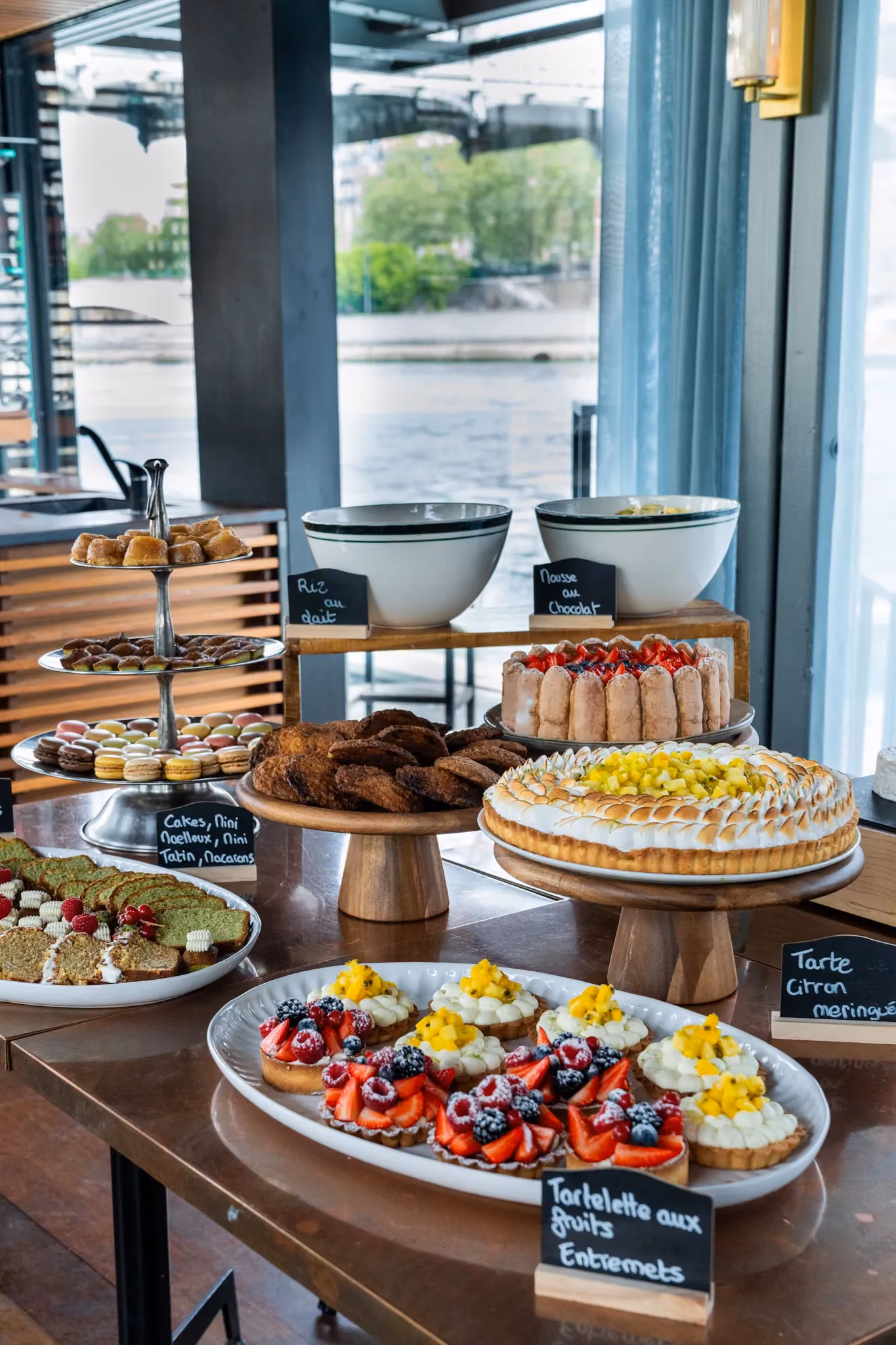 Table with assorted French desserts including tarts, macarons, cakes, and mousse by a riverside window.