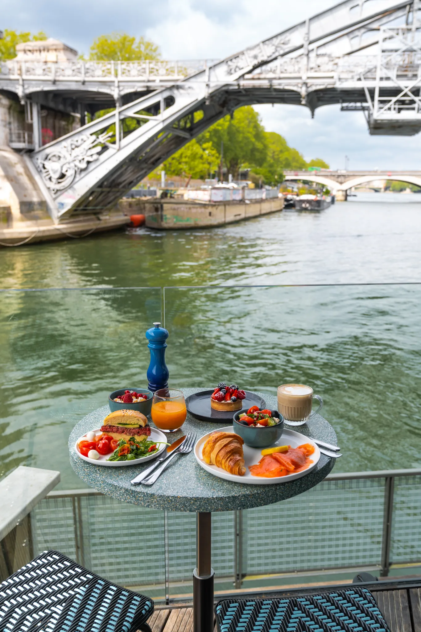 Table with breakfast including croissant, smoked salmon, salad, burger, juice, and coffee by river under a metal bridge.