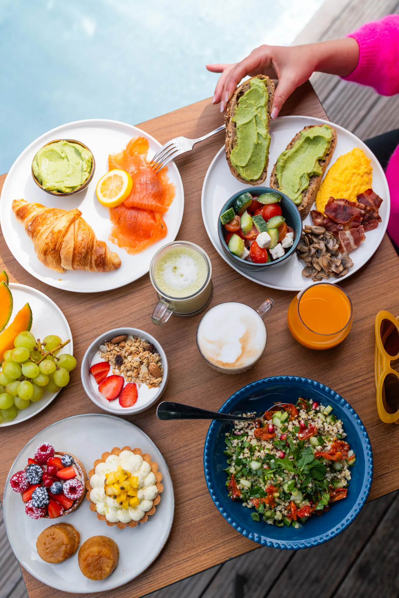 Brunch spread with avocado toast, scrambled eggs, croissant, salad, desserts, coffee, juice, and fruit on wooden table.