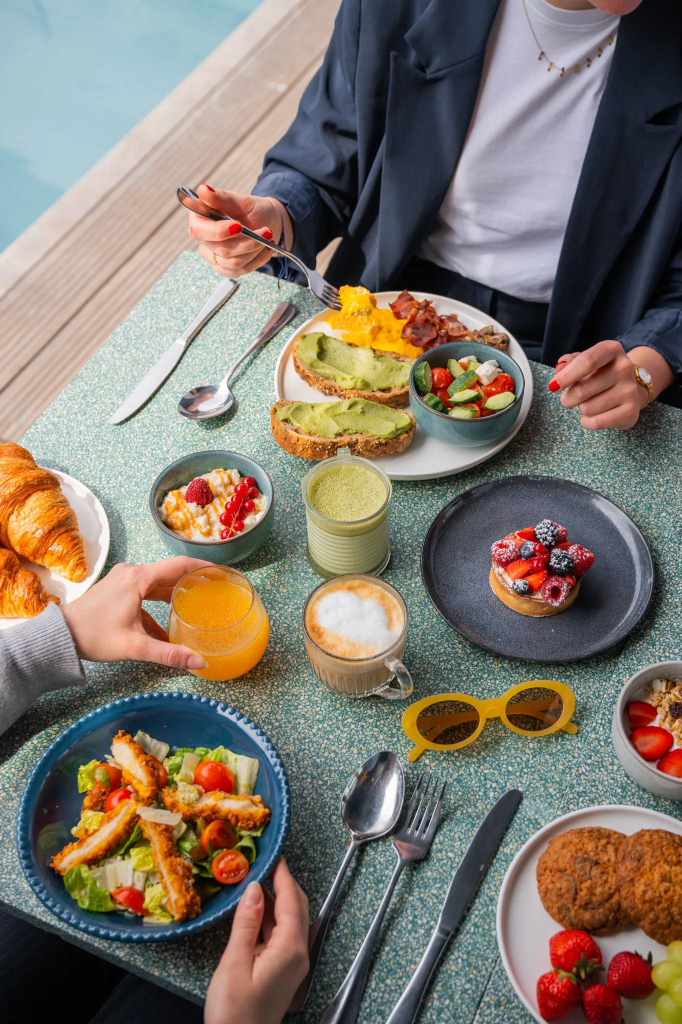 Two people enjoying a breakfast with avocado toast, salad, croissants, juice, coffee, and fruit dessert outdoors.