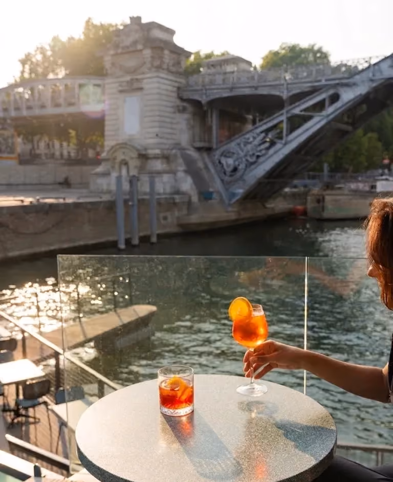 Person holding glass of orange cocktail by river with stone bridge in background at sunset.