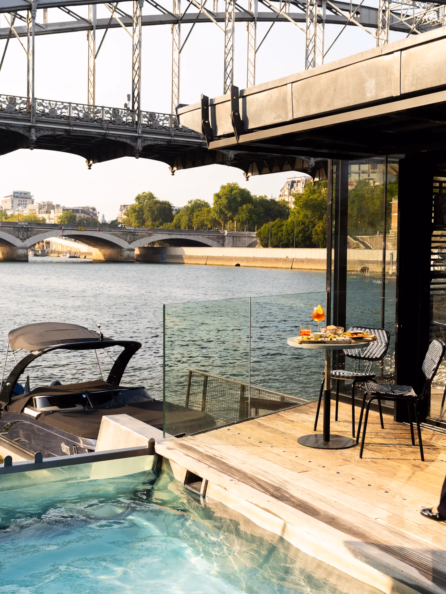 Outdoor table with food and drinks on a deck by a river with a boat and a bridge in the background.