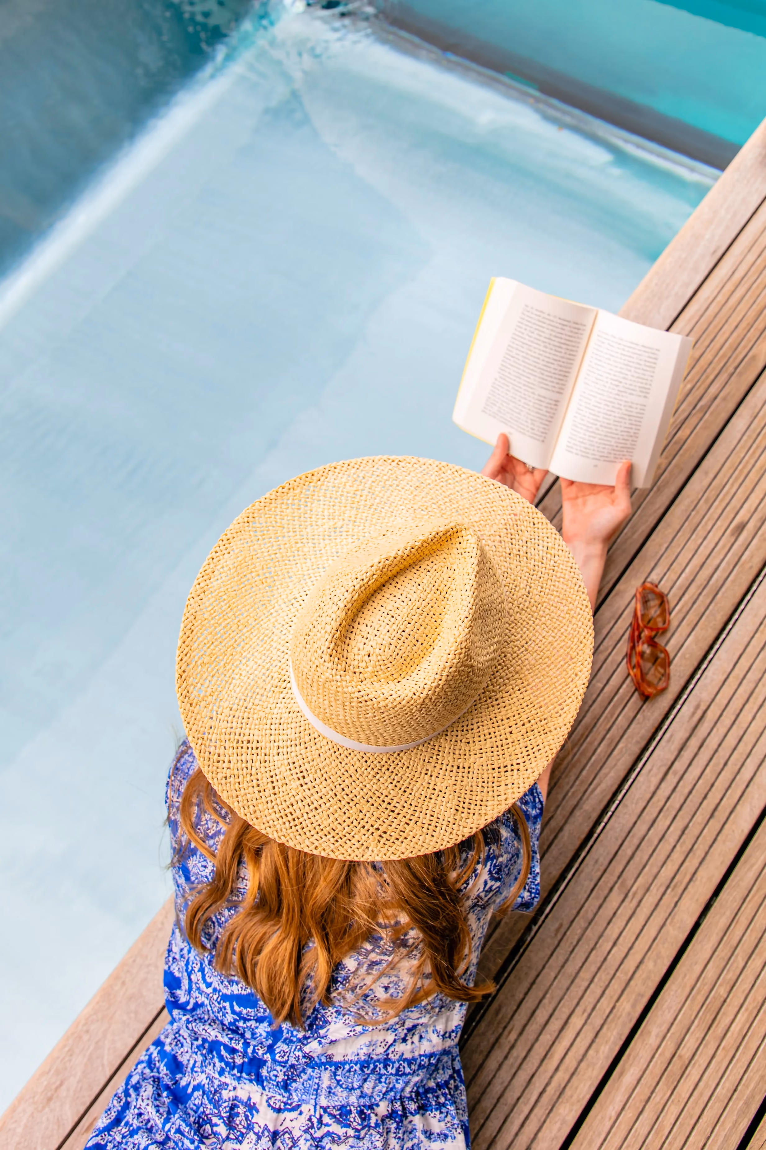 Person with long hair and a straw hat reading a book by a pool on wooden decking.