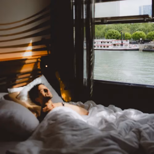 Man lying in bed stretching in sunlight near a window overlooking water and trees outside.