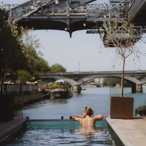 Person relaxing in a pool by a river with multiple bridges and greenery in the background.