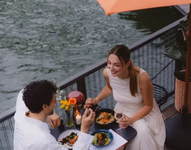 Couple dining at a table by the water, sharing food and drinks with flowers and candle on table.