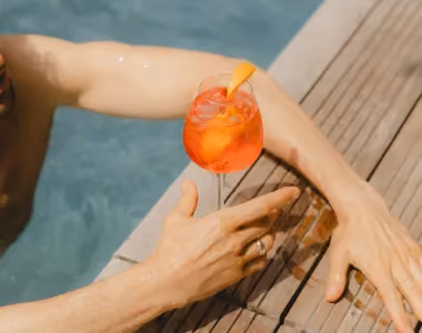 Person in pool holding a glass with an orange cocktail garnished with an orange slice on the edge.