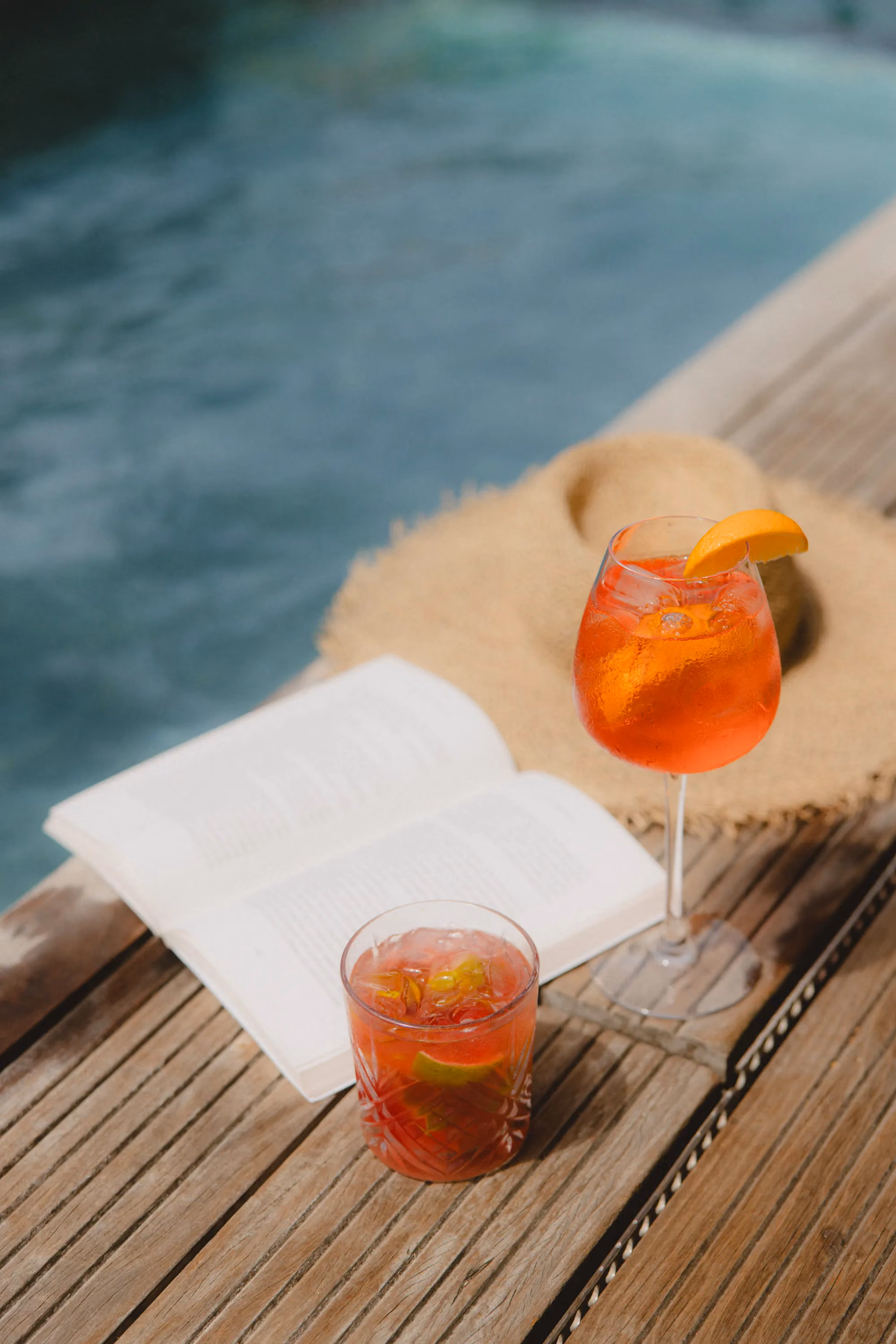 Two orange cocktails on wooden deck with open book and straw sunhat near a pool.