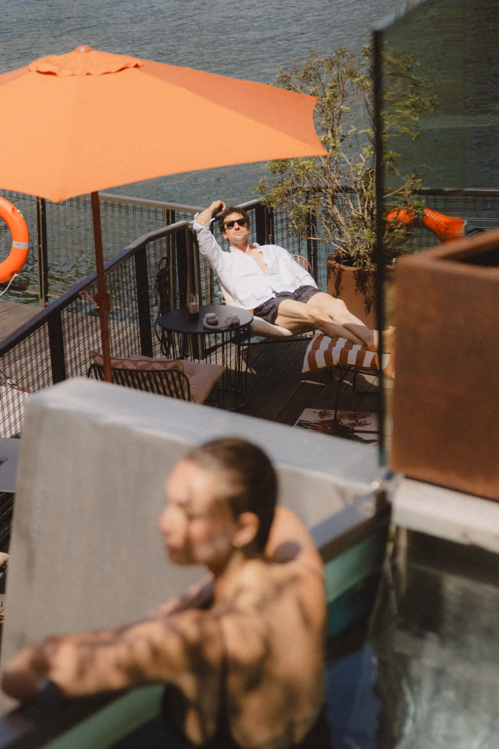 Man in white shirt and sunglasses relaxing on lounge chair under an orange umbrella by water.