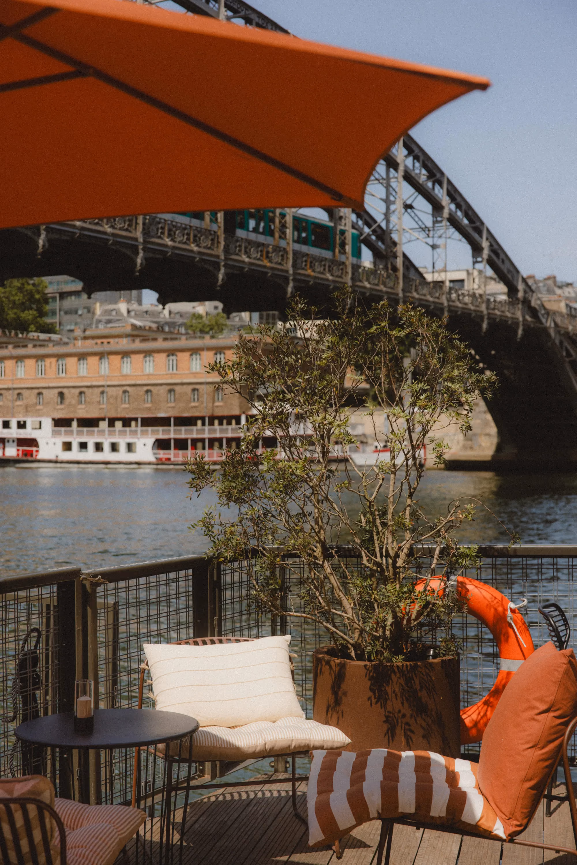 Outdoor seating with cushions under an orange umbrella by a river with a bridge and boat in the background.