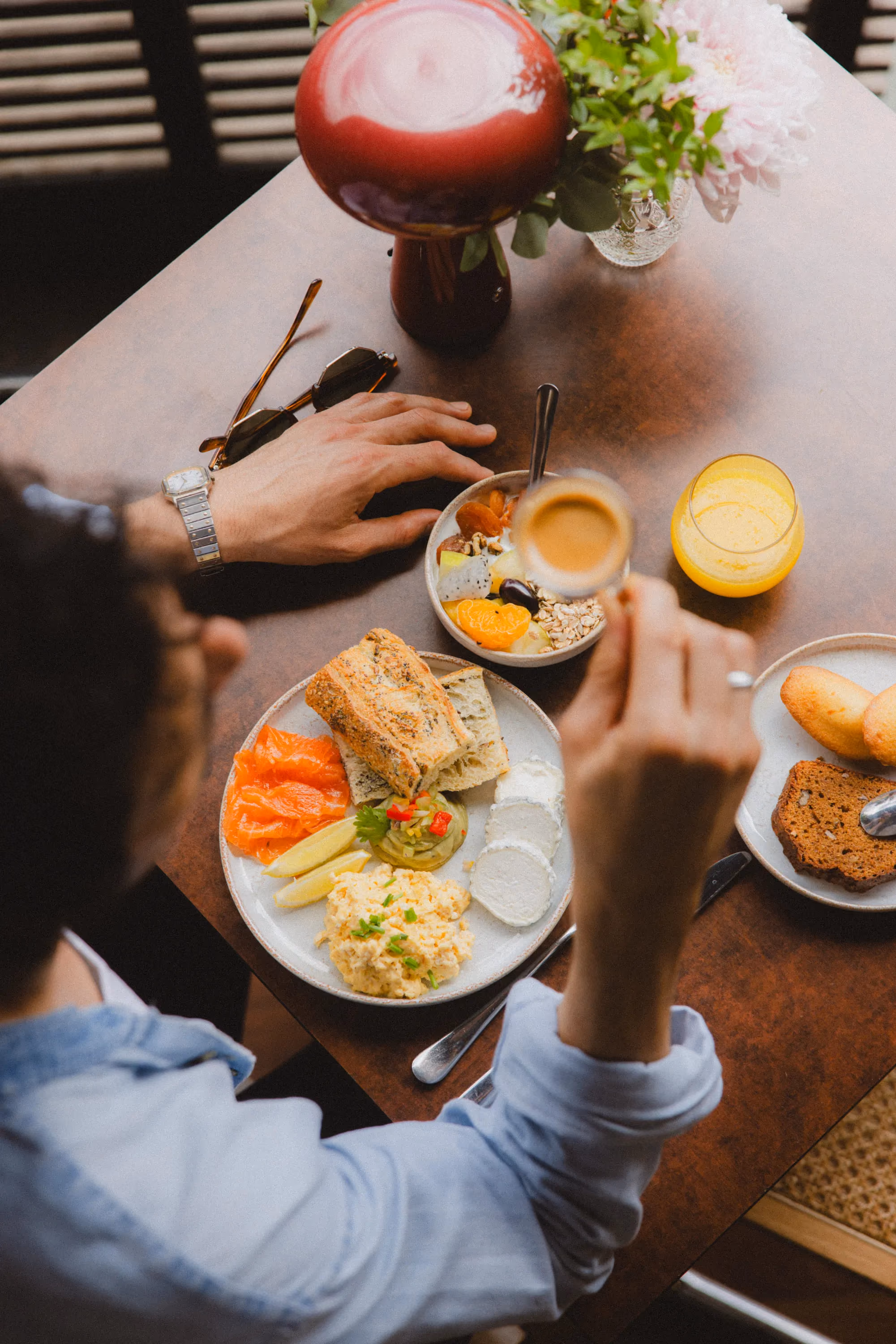Person holding a cup of coffee at a table with breakfast including bread, eggs, smoked salmon, cheese, fruit, and juice.