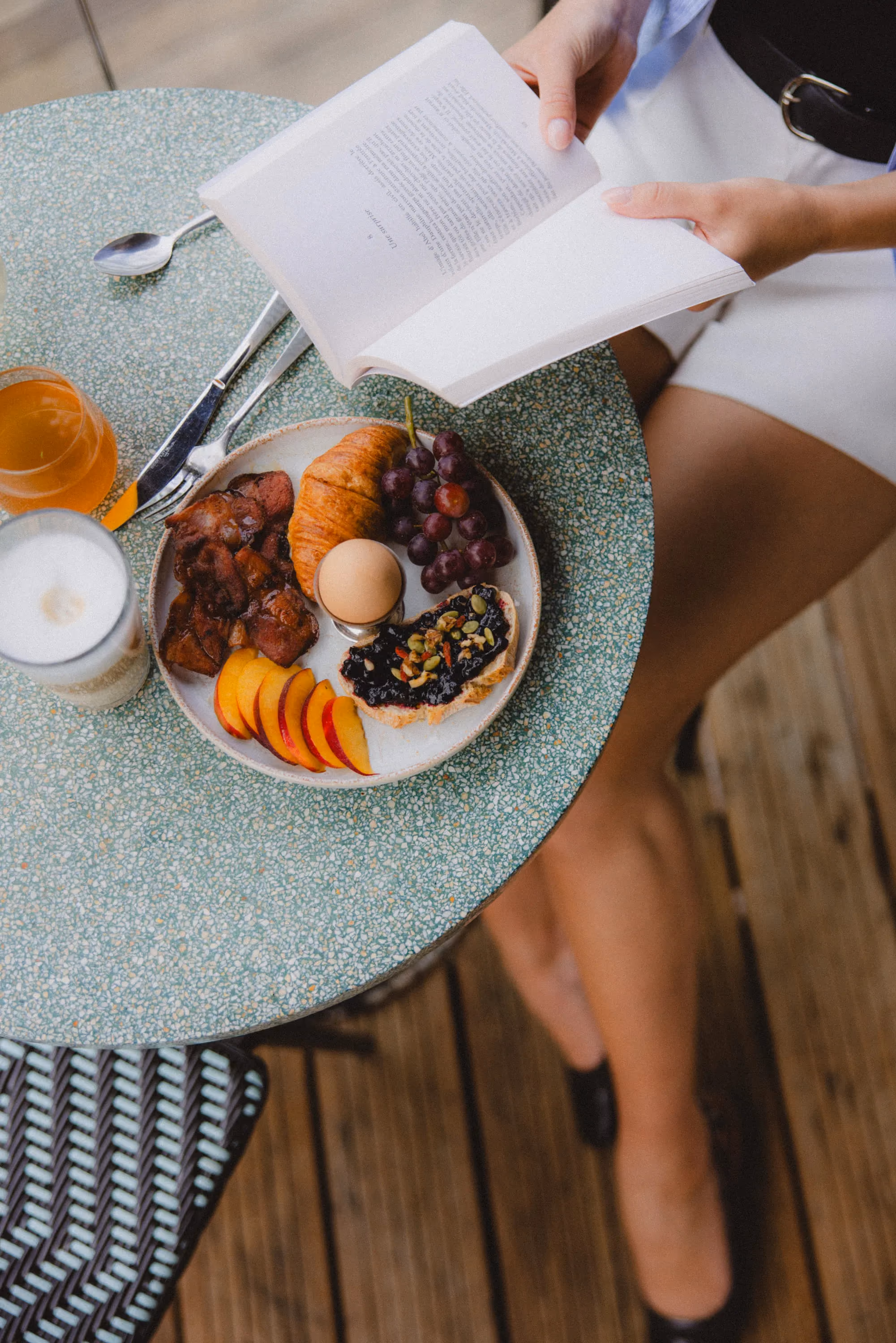 Person reading a book at a table with breakfast plate of bacon, croissant, egg, grapes, peach, and toast.