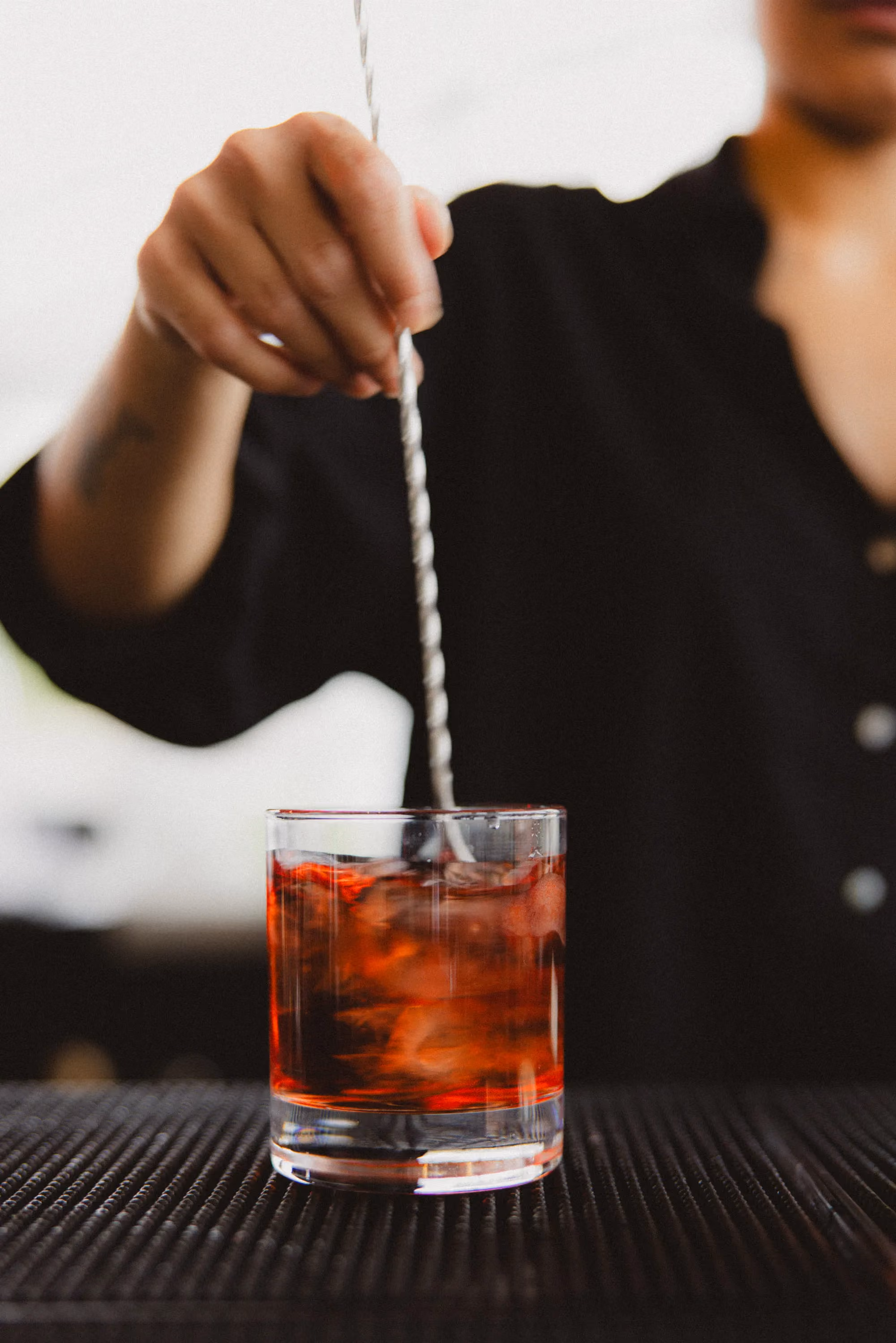 Person stirring a red cocktail with ice cubes in a short clear glass on a black mat.