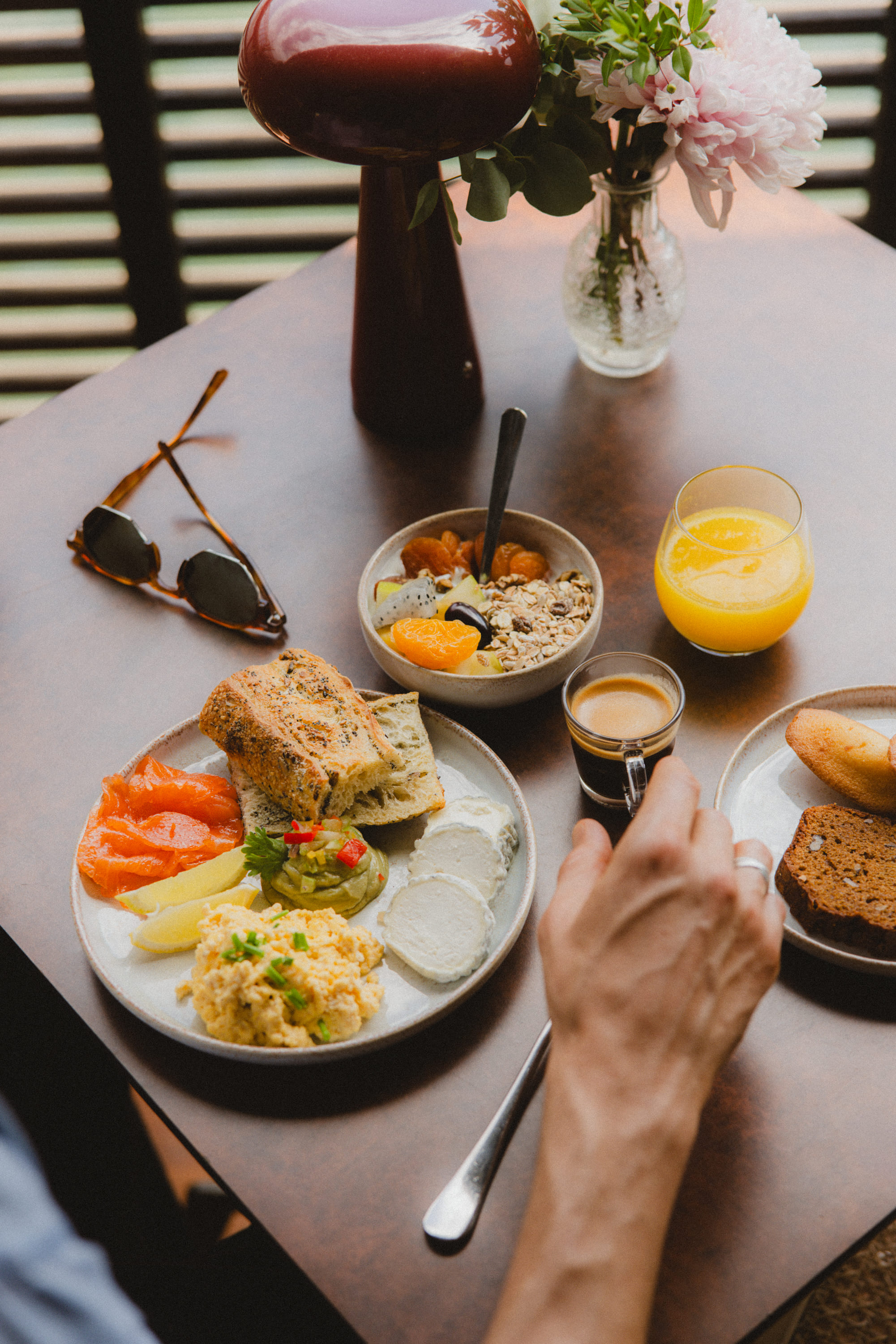 Person's hand reaching for espresso near plates with bread, scrambled eggs, cheese, fruit, muesli, and orange juice.