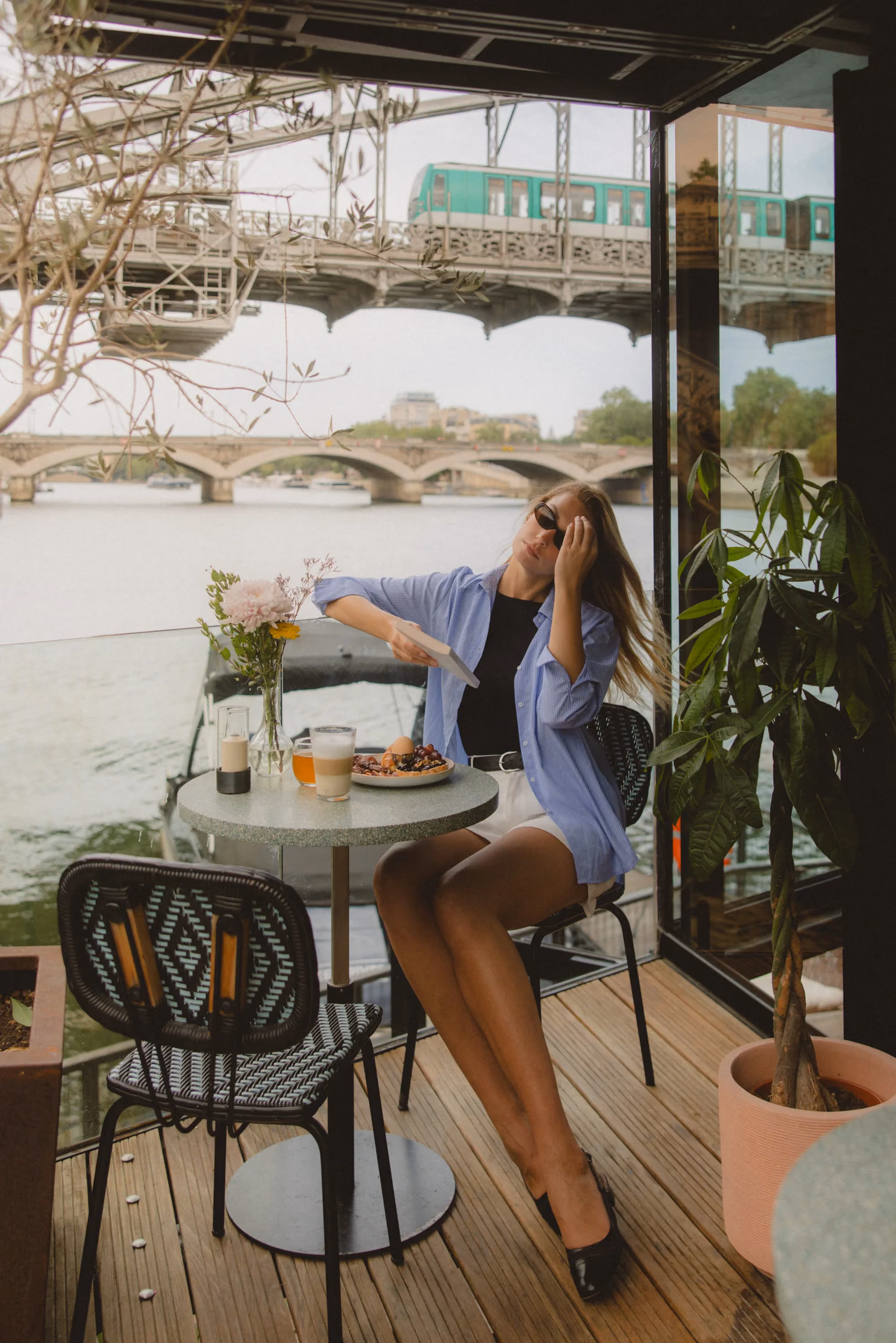 Woman in sunglasses sits at table with breakfast near river, bridge, and passing green train.