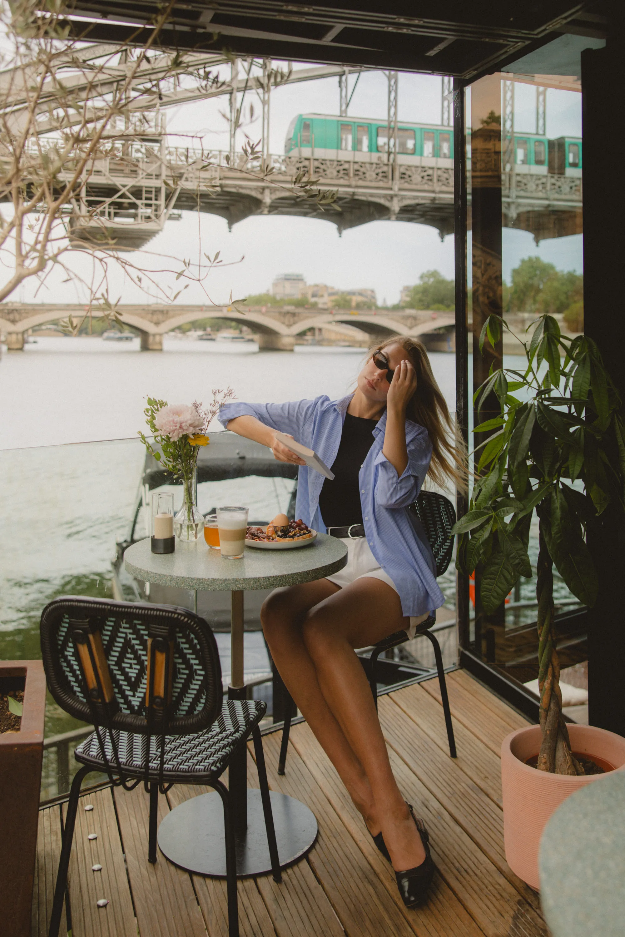 Woman in sunglasses sitting at a table by the river with food and drinks, a green train crosses bridge above.