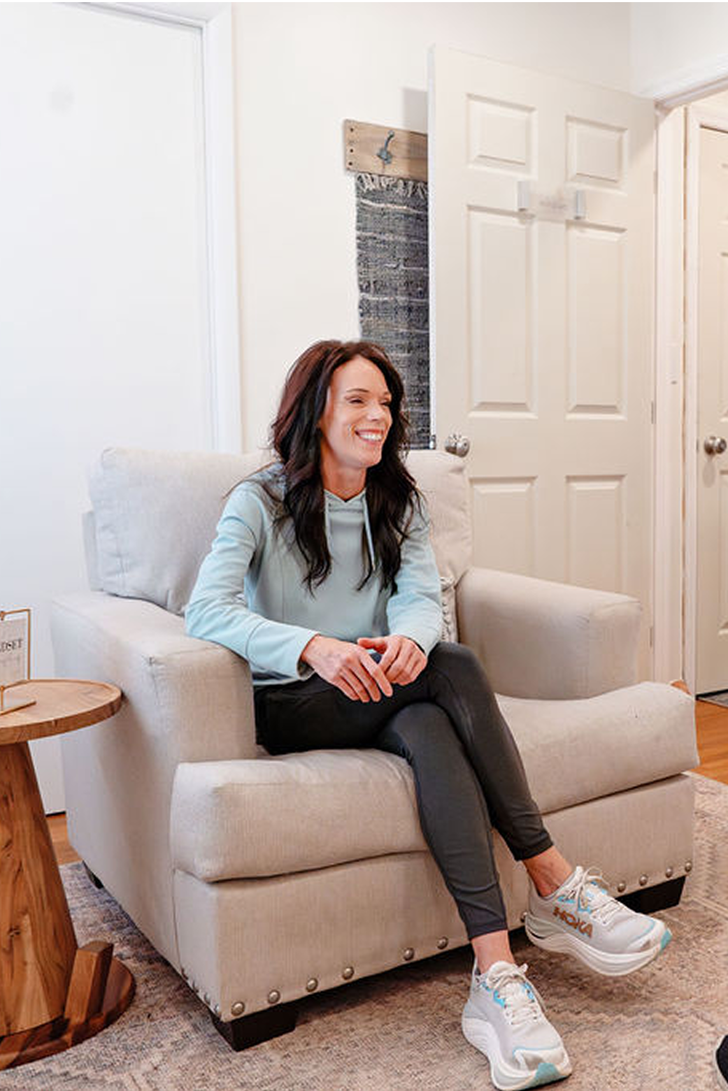 Woman with long dark hair wearing a light blue hoodie, dark pants, and white sneakers sitting and smiling on a beige armchair in a living room.