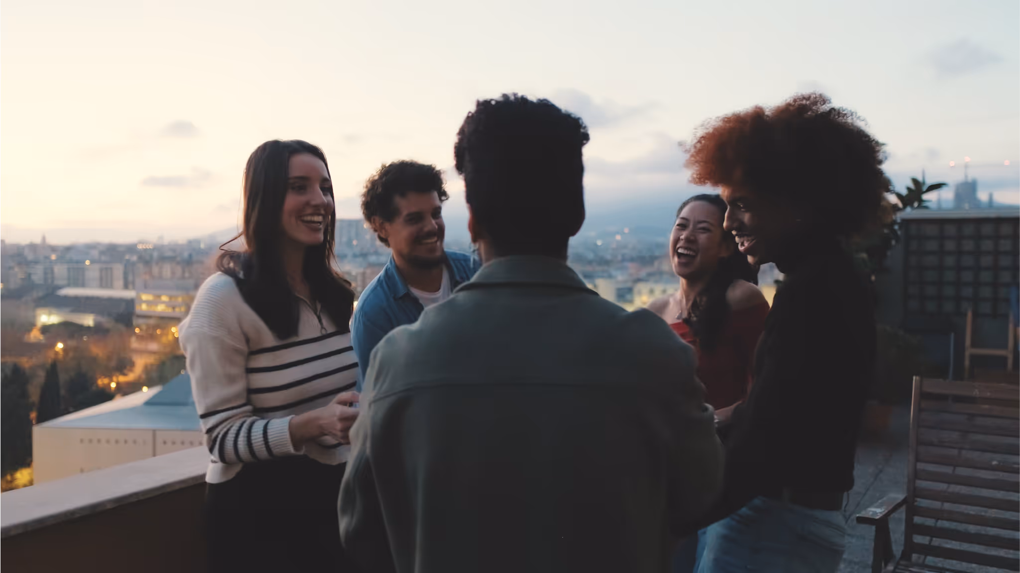 Group of five young adults laughing and socializing on a rooftop terrace at dusk with cityscape in the background.