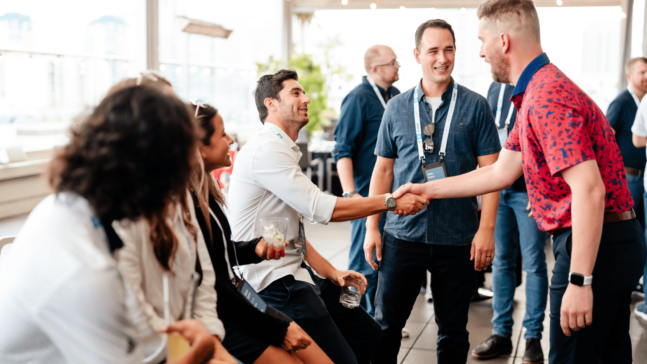 Two men shaking hands at a casual networking event with other attendees smiling and conversing in the background.
