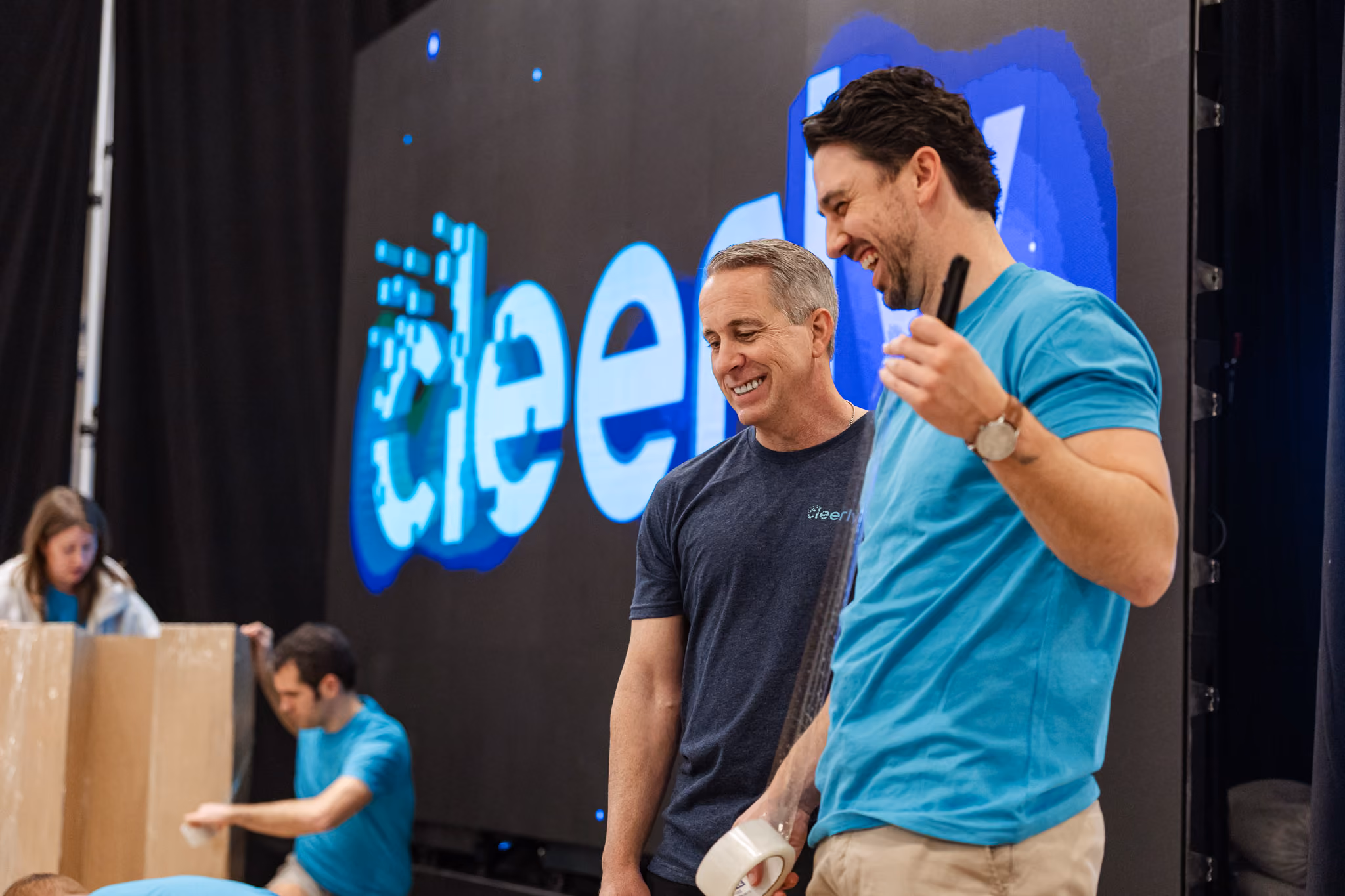 Two men smiling and talking in front of a large screen with 'cleerly' logo during setup.